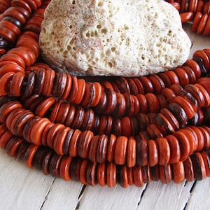 Può includere: Una collana di perline di legno di colore arancione e marrone. Le perline sono rotonde e piatte, e sono disposte a spirale. Le perline sono infilate su un filo, e sono appoggiate su una superficie bianca.