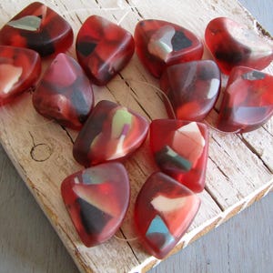 Op de afbeelding: Een verzameling doorschijnende rode, onregelmatig gevormde kralen met ingebedde kleurrijke fragmenten. De kralen zijn geregen en liggen op een verweerd houten oppervlak, met een mix van rode, witte, zwarte en groene tinten.