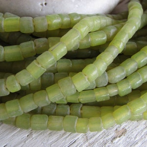 以下が含まれることがあります： 明るい緑色の長方形のビーズの山をクローズアップ。ビーズは複数本のストランドを形成するように一緒に紐で繋がれています。ビーズはわずかに半透明で、色は一貫しています。ビーズはおそらくジュエリー作りのためです。