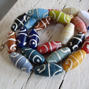 Può includere: Una collezione di perline ovali colorate con disegni a spirale bianchi. Le perline sono in vari colori, tra cui blu, rosso, giallo e nero. Sono disposte su una superficie di legno bianco e invecchiato, suggerendo un tema artigianale o di gioielleria.