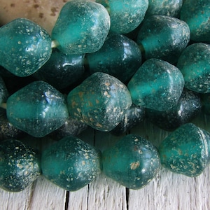 Pode incluir: Um fio de contas de vidro azul turquesa com uma superfície texturizada e salpicada. As contas são facetadas e têm uma forma ligeiramente irregular.