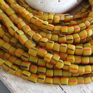 Può includere: Una collana di perline di vetro gialle con strisce nere e arancioni. Le perline sono quadrate e hanno una finitura lucida.