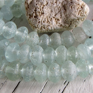 Op de afbeelding: Close-up van lichtblauwe, matte glazen kralen met een gespikkeld uiterlijk. De kralen zijn ovaal en aan elkaar geregen. Een poreuze, beige steen ligt achter de kralen. Ideaal voor het maken van sieraden.