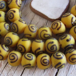 May include: Oval-shaped yellow beads with dark brown spiral patterns. The beads have a translucent quality and are scattered on a light wooden surface. These beads are suitable for jewellery making and other craft projects.