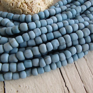 Può includere: Primo piano di una collezione di perline azzurre a forma di botte. Le perline sono infilate insieme, creando un effetto strutturato e stratificato. Le perline hanno una finitura opaca e sono disposte su una superficie di legno chiaro.
