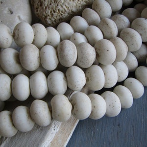 Pode incluir: Um fio de contas redondas de cerâmica de cor branca com textura salpicada. As contas são enfiadas em um cordão branco.