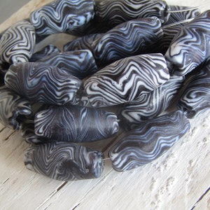 Op de afbeelding: Een verzameling zwarte en witte gemarmerde glazen kralen. De kralen zijn ovaalvormig en hebben een draaiend patroon.