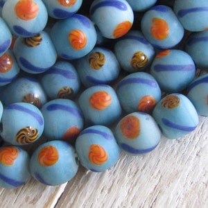 Könnte beinhalten: Eine Sammlung blauer Glasperlen mit orangefarbenen und braunen Wirbelmustern. Die Perlen haben eine matte Oberfläche und sind aufgereiht.