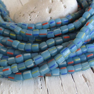 May include: A strand of light blue glass beads with white, red, and yellow stripes. The beads are square shaped and have a frosted finish.