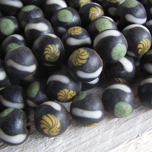 Peut inclure: Gros plan sur un ensemble de perles sombres avec des rayures blanches et des motifs décoratifs en spirale jaunes et verts. Les perles sont rondes et semblent être en verre ou dans un matériau similaire. Ces perles sont probablement destinées à la fabrication de bijoux.