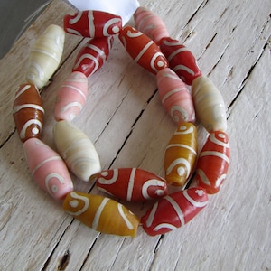 Può includere: Una collana composta da perline ovali in vari colori, tra cui rosso, rosa, giallo e crema. Ogni perla presenta un disegno bianco a spirale. Le perline sono infilate insieme, creando una forma circolare. La collana è su legno.