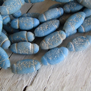 以下が含まれることがあります： 淡い青色の樽型のビーズのコレクション。風化したテクスチャード加工が施されています。ビーズはわずかに不規則な形状をしており、紐で繋がれていることから、ジュエリー作りや手芸用であることがわかります。明るい色の木製の表面にビーズが展示されています。