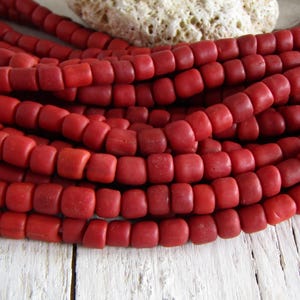Op de afbeelding: Close-up van strengen rode, vierkante kralen. De kralen hebben een diepe, matte rode kleur en lijken van een natuurlijk materiaal te zijn gemaakt. De kralen zijn gerangschikt in meerdere strengen, waardoor een getextureerd, gelaagd effect ontstaat. Op de achtergrond bevindt zich een wit, getextureerd object.