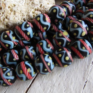 Op de afbeelding: Een streng zwarte glazen kralen met een kleurrijk geometrisch patroon. De kralen zijn versierd met rode, blauwe en gele strepen en stippen.