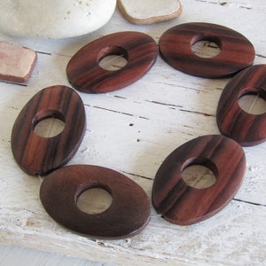 Può includere: Un set di sei perline di legno di forma ovale con venature di legno marrone scuro e un foro al centro. Le perline sono infilate su un filo bianco.