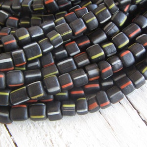 Può includere: Primo piano di una collezione di perline scure e quadrate con strisce colorate. Le perline sono principalmente nere con strisce bianche, gialle e rosse. Le perline sono disposte in un motivo circolare, suggerendo una collana o un braccialetto.