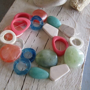 Può includere: Una collezione di perline colorate e testurizzate, tra cui rosa, verde, blu e bianco. Le perline sono disposte in un cerchio su una superficie di legno bianco.