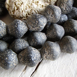 Könnte beinhalten: Eine Kette aus dunkelgrauen, unregelmäßig geformten Perlen mit einer rauen, strukturierten Oberfläche. Die Perlen sind aus Naturstein und haben ein rustikales, erdiges Aussehen.