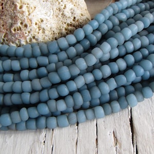 Può includere: Primo piano di più fili di perline azzurre. Le perline hanno una finitura opaca e sono piccole e rotonde. Un pezzo di pietra beige e porosa è sullo sfondo. Le perline sono esposte su una superficie di legno bianco e invecchiato.