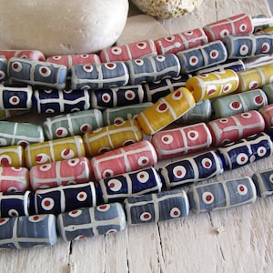 Può includere: Una collezione di perline di vetro colorate con motivi geometrici. Le perline sono cilindriche e hanno accenti bianchi, blu, rossi, gialli e rosa.