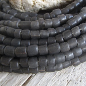 Op de afbeelding: Een streng donkergrijze glazen kralen, elke kraal is cilindrisch en licht afgerond. De kralen zijn in een close-up weergegeven, waardoor de textuur en kleur van het glas zichtbaar zijn.