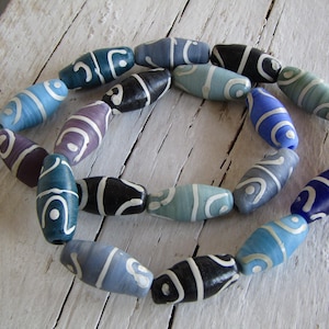 Può includere: Una collana di perline ovali colorate. Le perline sono dipinte in tonalità di blu, viola e nero, con disegni a spirale bianchi. Le perline sono infilate insieme, creando una forma circolare. Lo sfondo è una superficie di legno bianco invecchiato.