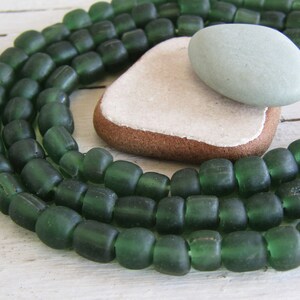 May include: Dark green glass beads with a rectangular shape, strung together. A smooth, oval, light green stone rests on a piece of textured, off-white paper and a brown, rounded stone. The beads and stones are arranged on a white surface.