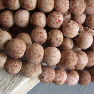 Könnte beinhalten: Eine Strähne aus beige und rot gesprenkelten Perlen. Die Perlen sind rund und haben eine raue, strukturierte Oberfläche.