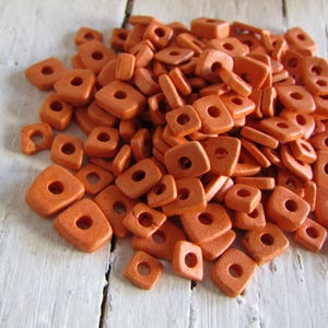 Può includere: Un mucchio di perline quadrate di terracotta arancione con un foro al centro di ogni perlina. Le perline sono disposte in uno schema casuale e sono tutte della stessa dimensione.