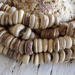 May include: A close-up shot of a necklace composed of light brown, cylindrical beads with darker brown markings. The beads are threaded together, forming layers. A textured, off-white stone is visible in the background.