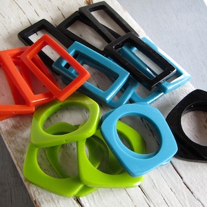 Può includere: Una collezione di componenti per gioielli in resina quadrati e rettangolari colorati. I componenti sono nelle tonalità di arancione, verde, blu e nero. Le forme sono impilate su una superficie di legno bianco.