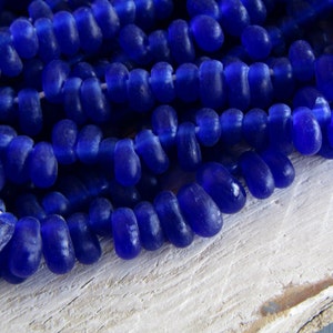 Puede incluir: Una hebra de cuentas de vidrio azul oscuro, cada cuenta es ligeramente irregular en forma y tiene un acabado mate.