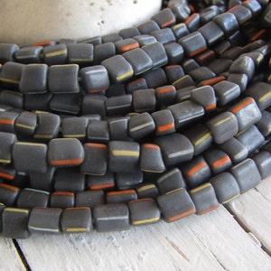 Può includere: Perle di vetro quadrate nere con strisce bianche e arancioni. Le perle sono impilate in un mucchio.