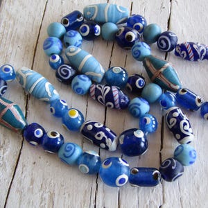 May include: A necklace with blue glass beads in various shades. The beads have patterns like swirls, dots, and stripes. The necklace is displayed on a white wooden surface. The beads are in shades of light, medium, and dark blue.
