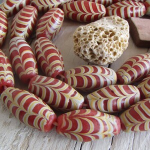 Op de afbeelding: Een streng rode en witte glazen kralen met een draaiende patroon. De kralen zijn ovaalvormig en hebben een glanzende afwerking.