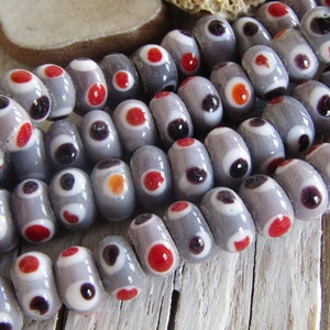 Op de afbeelding: Grijze glazen kralen met witte en rode cirkels. De kralen hebben een gespikkeld, getextureerd oppervlak.