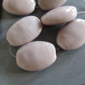 Può includere: Sei perline ovali color malva chiaro infilate. Ogni perlina ha una superficie liscia e lucida con motivi delicati. Sono infilate su un filo sottile e trasparente, probabilmente per gioielli o progetti artigianali. Ogni perlina misura circa 2 cm di lunghezza.