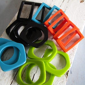 Può includere: Una collezione di componenti per gioielli in resina colorata e geometrica in varie forme. L'assortimento comprende pezzi neri, turchesi, arancioni e verdi, ideali per la creazione di bracciali, collane o orecchini. I componenti sono disposti su una superficie di legno invecchiato.