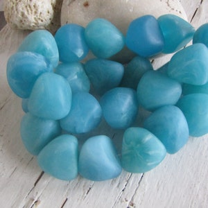 Può includere: Un gruppo di perline lisce e arrotondate, di colore azzurro. Le perline hanno un aspetto leggermente traslucido e variano nelle tonalità di blu. Sono disposte insieme, suggerendo che facciano parte di un progetto di gioielleria o artigianato.