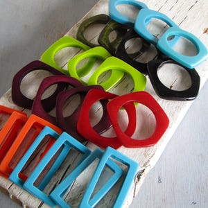 Può includere: Una collezione di braccialetti in resina colorati e geometrici in varie tonalità, tra cui arancione, rosso, viola, verde, nero e blu. I braccialetti sono disposti su una superficie di legno bianco, mostrando i loro colori vivaci e il design moderno.