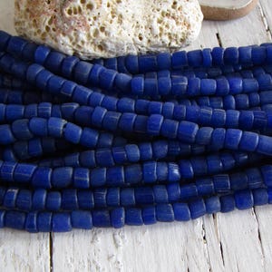 May include: A strand of deep blue glass beads, each bead is square shaped with rounded edges. The beads are arranged in a pile, showing the texture and colour of the glass.