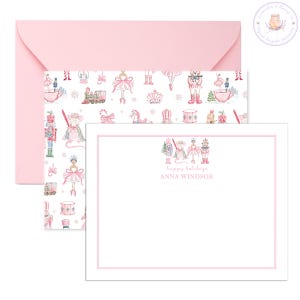 May include: A stationery set featuring a pink envelope, a patterned card, and a blank card with a pink border. The patterned card has a repeating design of ballet dancers, nutcrackers, and other holiday-themed illustrations. The blank card has the text "happy holidays" and "ANNA WINDSOR".