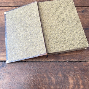 May include: A hardcover book with a gold and black damask patterned cover. The book is partially open, revealing the front cover and the spine area. The pattern consists of intricate, repeating floral and scroll motifs in black on a gold background.