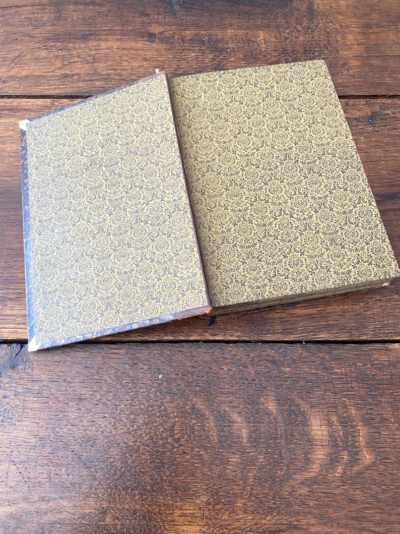 May include: A hardcover book with a gold and black damask patterned cover. The book is partially open, revealing the front cover and the spine area. The pattern consists of intricate, repeating floral and scroll motifs in black on a gold background.