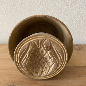 May include: A wooden, round, carved butter mold with a pineapple design. The mold is in two pieces, with the top piece sitting inside the bottom piece.