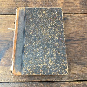 May include: An antique hardcover book featuring a black leather spine and a marbled cover with a mottled pattern of black and gold. The book shows signs of wear along the edges and the spine, with visible fraying and exposed material on the binding.
