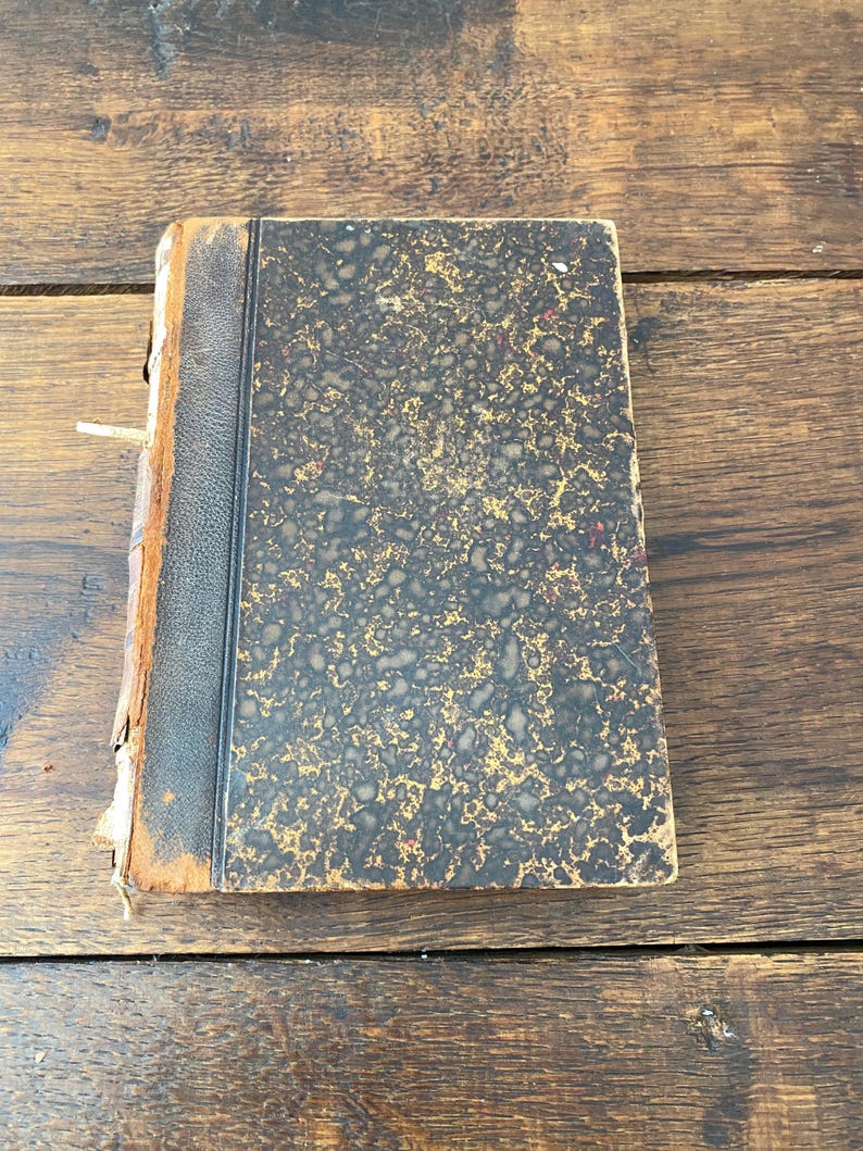 May include: An antique hardcover book featuring a black leather spine and a marbled cover with a mottled pattern of black and gold. The book shows signs of wear along the edges and the spine, with visible fraying and exposed material on the binding.