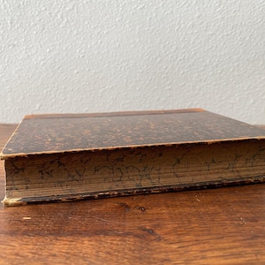 May include: A vintage hardcover book featuring a dark brown cover with a mottled, speckled pattern. The edges of the pages are decorated with a dark, marbled design that matches the aesthetic of the cover, showing signs of age and wear.