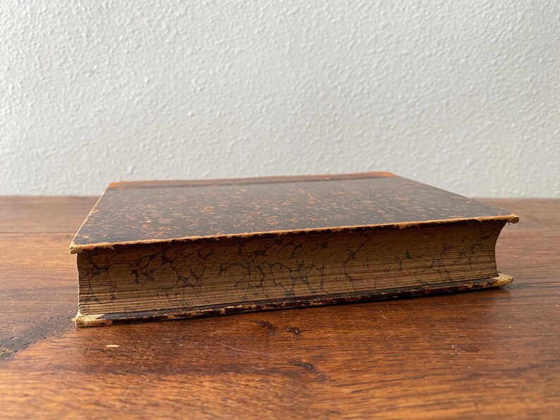 May include: A vintage hardcover book featuring a dark brown cover with a mottled, speckled pattern. The edges of the pages are decorated with a dark, marbled design that matches the aesthetic of the cover, showing signs of age and wear.