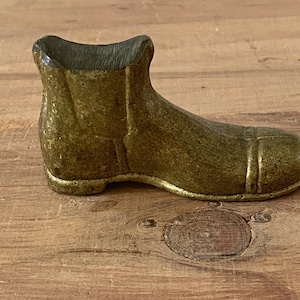 May include: A small, antique-style boot figurine in a metallic gold color. The boot has a rounded toe and a short, open top. The surface has a textured appearance, suggesting an aged or weathered finish. The boot is resting on a wooden surface.